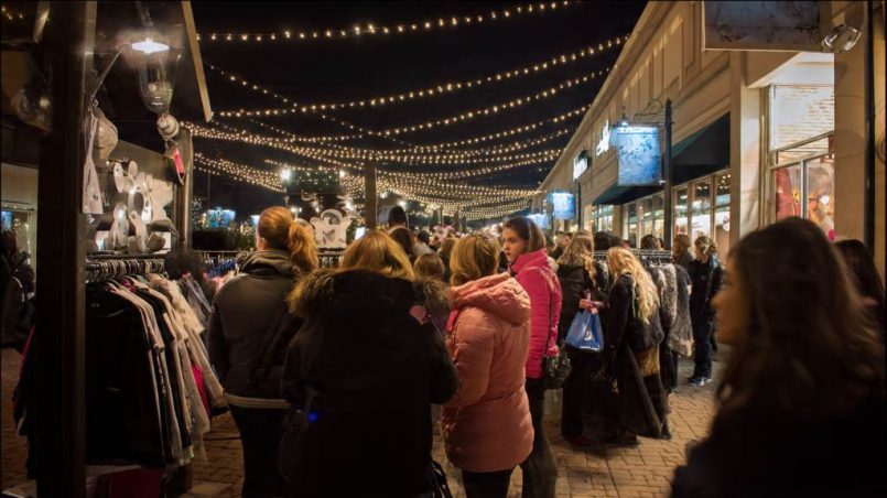 Shoppers excitedly browsing racks at a bustling Black Friday outlet mall entrance under festive lights