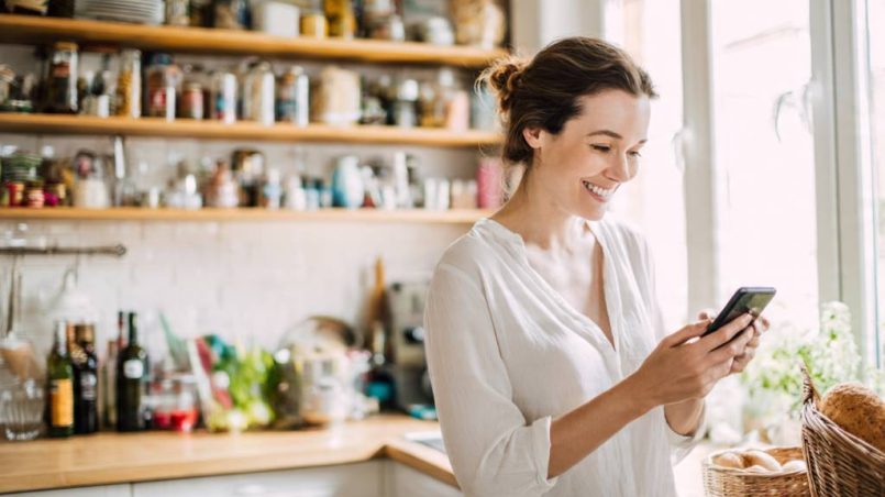 Smiling mom checking grocery list on phone in bright kitchen with full pantry